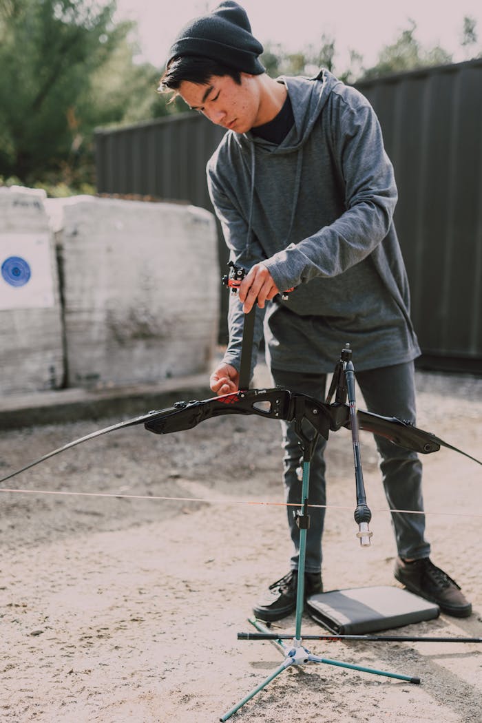 Young archer adjusting a compound bow at an outdoor archery range during the day.