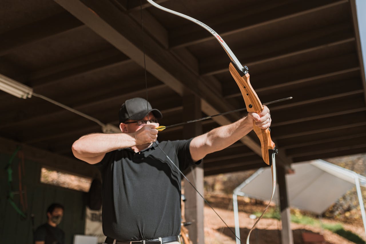 An adult male archer aiming his traditional bow outdoors at an archery range.