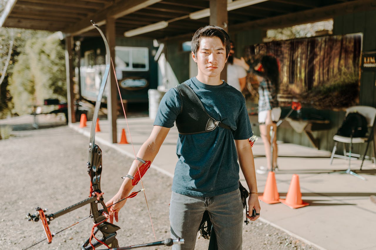 Home A young adult man practicing archery at an outdoor range on a sunny day.