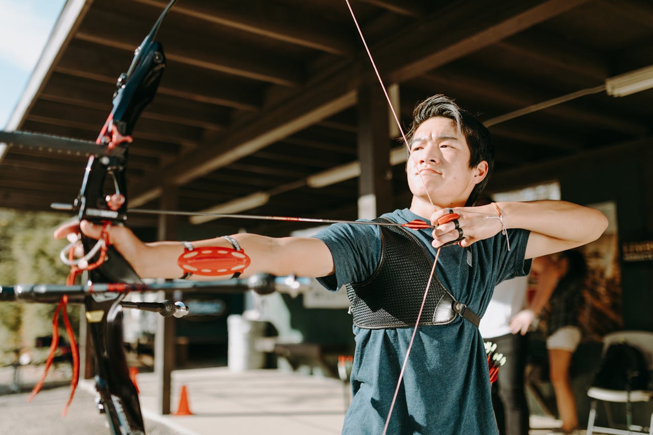 Home An adult male archer intently aims a recurve bow at an outdoor archery range.