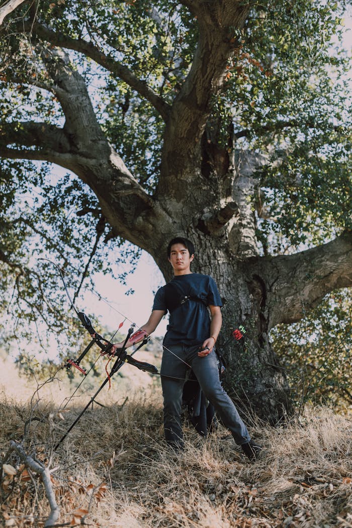 Home Young archer in nature practicing archery with a compound bow under a large tree.